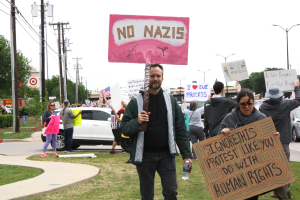 A protester holds a pink sign reading “NO NAZIS” while a woman next to him holds a sign that says “IGNORE THIS PROTEST LIKE YOU DO WITH HUMAN RIGHTS,” calling attention to ongoing human rights concerns.